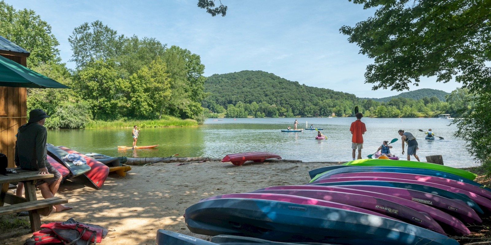 A scenic lakeside area with stacked kayaks, people kayaking, and a lush green backdrop of trees and hills under a clear blue sky.