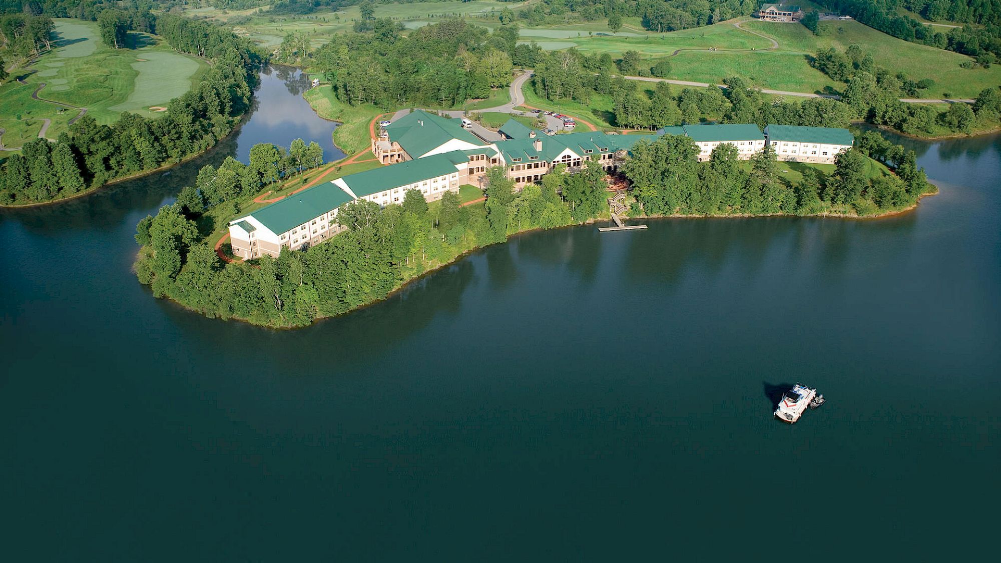 An aerial view of a large building complex surrounded by water on three sides, with lush green trees and a small boat in the water nearby.