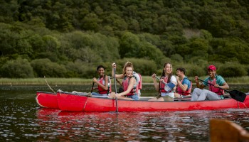 A group of five people wearing life jackets are paddling in a red canoe on a body of water surrounded by lush greenery.