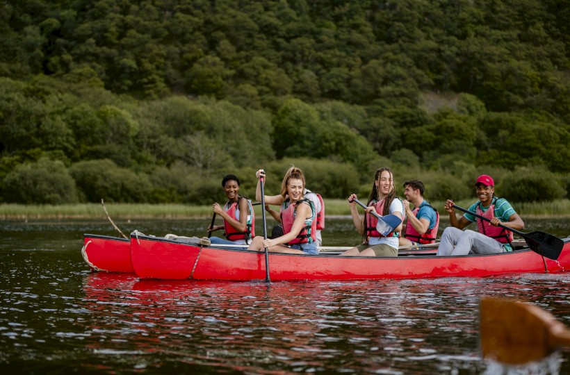 A group of five people wearing life jackets are paddling in a red canoe on a body of water surrounded by lush greenery.