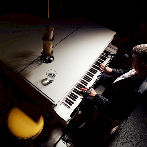 A person is playing a white grand piano, with a glass and two candles on top, in a dimly lit setting.