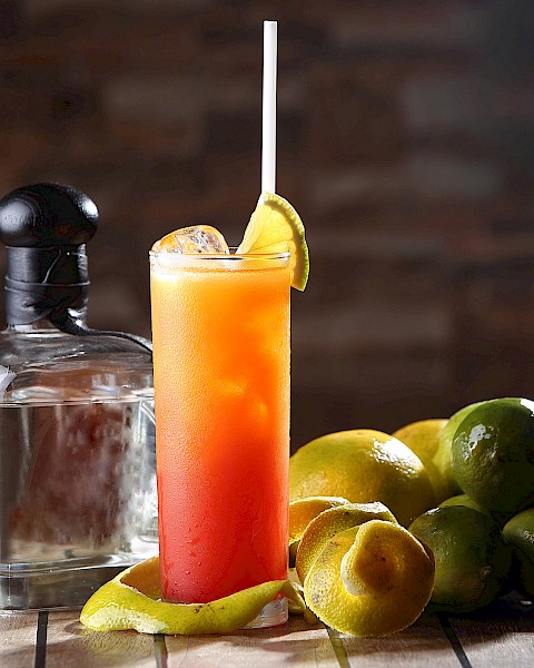 A cocktail with a straw and lime garnish, surrounded by limes and a liquor bottle on a wooden table.