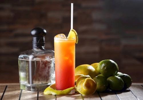 A cocktail with a straw and lime garnish, surrounded by limes and a liquor bottle on a wooden table.