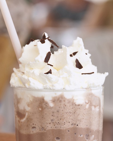 A chocolate frozen drink topped with whipped cream and chocolate shavings, served in a glass with a straw.
