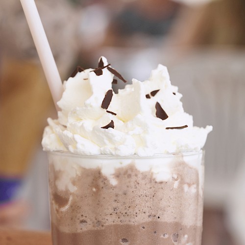 A chocolate frozen drink topped with whipped cream and chocolate shavings, served in a glass with a straw.