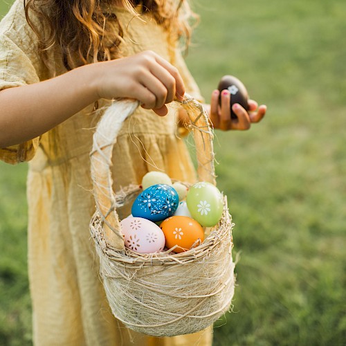 A child in a yellow dress holds a basket filled with colorful, decorated eggs, standing on grass.