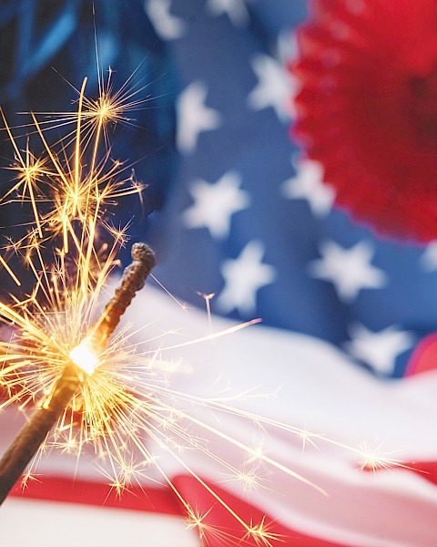 A lit sparkler in front of an American flag and festive decorations, creating a celebratory and patriotic scene.