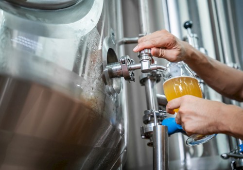 Someone filling a glass with beer from a large stainless steel brewing tank in a brewery setting.