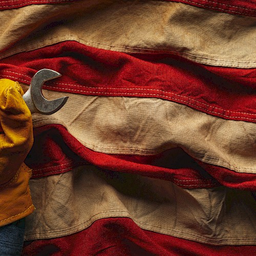 A gloved hand holding wrenches emerges from a wrinkled American flag, symbolizing labor and patriotism.