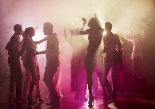 A group of people dancing in a dimly lit, foggy environment with colorful lights and silhouettes visible.