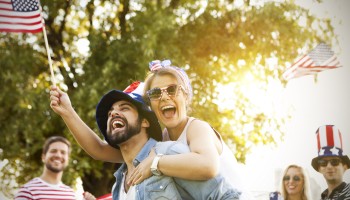 A group celebrates outdoors with American flags, wearing patriotic outfits and accessories, as one person gets a piggyback ride.