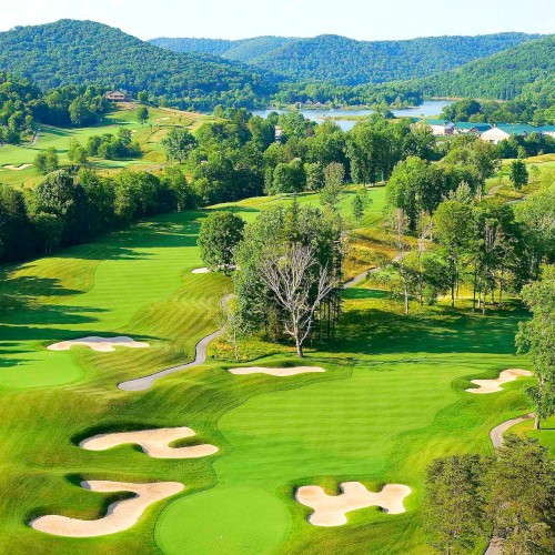 A lush golf course with sand bunkers and manicured greens, set against a backdrop of rolling hills and a distant water body.