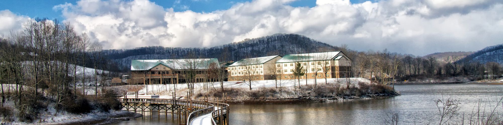 A serene landscape with buildings by the water, a curved snowy walkway leading to them, surrounded by mountains and a partly cloudy sky.