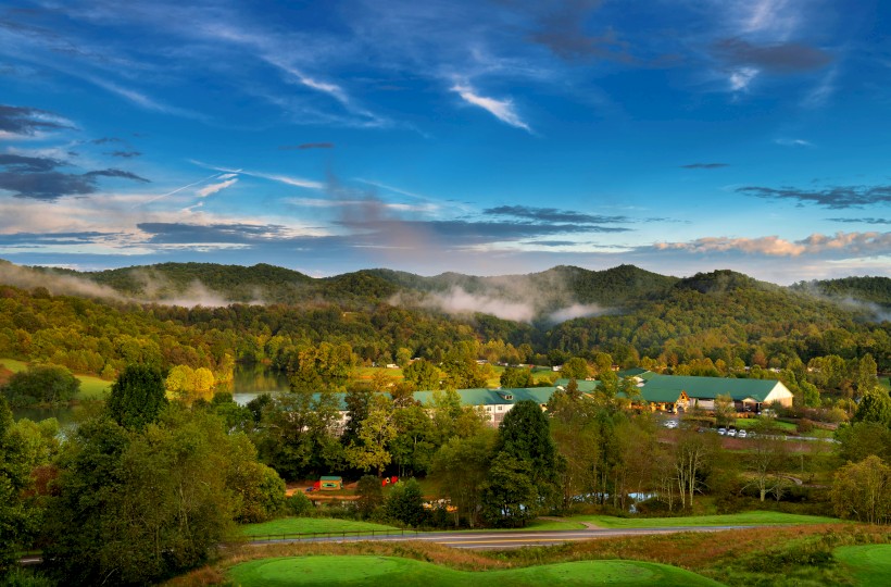 A picturesque landscape featuring rolling hills, a forest, and buildings with green roofs under a vivid blue sky scattered with clouds.