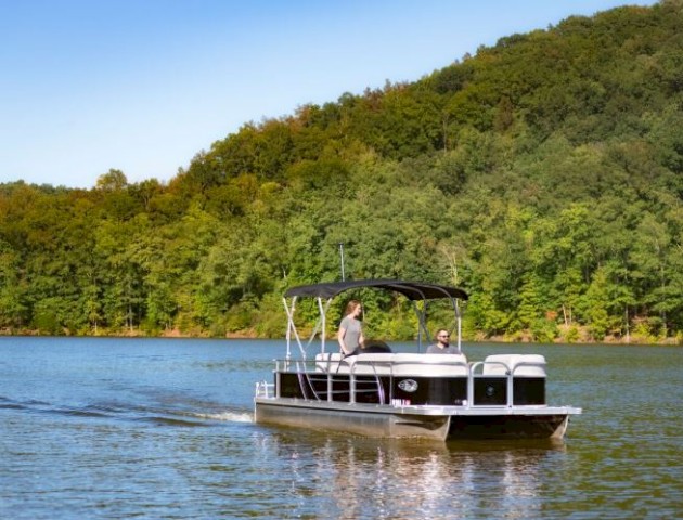 A pontoon boat with two people is on a calm lake with a forested hillside in the background under a clear blue sky.