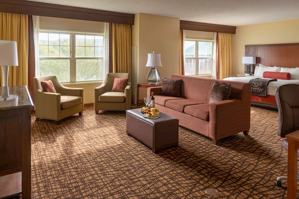 A hotel room features a bed, sofa, two chairs, a coffee table with wine, lamps, and large windows with curtains, offering a cozy atmosphere.