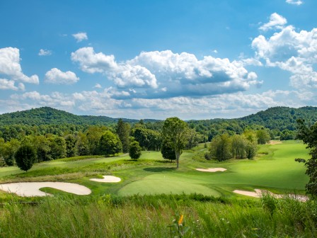 A scenic view of a golf course with manicured grass, sand bunkers, trees, and distant hills under a partly cloudy sky.