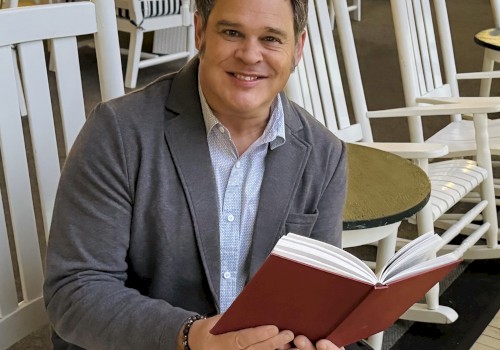 A person is sitting in a white rocking chair, holding an open red book, in an area with several chairs and large windows.