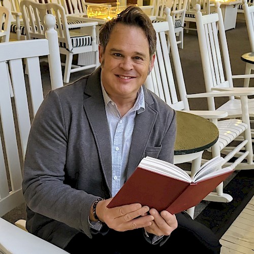A person is sitting in a chair, smiling, while holding an open book in a setting with several similar chairs around.