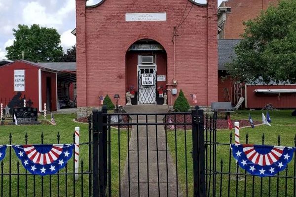 A red brick building with a gated entrance, decorated with patriotic banners, situated in a grassy yard with trees on a cloudy day.