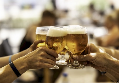 Four people clinking glasses of beer in a celebratory toast, with a blurred background indicating an indoor social gathering.