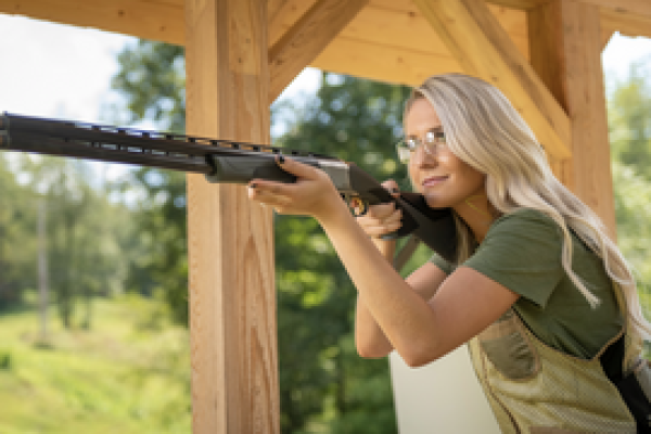 Blonde woman aims a shotgun awaiting a clay pigeon to be released while standing under a pavilion.