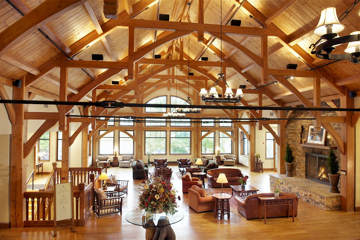A spacious room with wooden beams, cozy seating, a large fireplace, and chandeliers, with abundant natural light coming from arched windows.