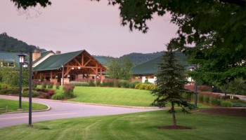 A scenic view shows a wooden building with a green roof, surrounded by lush grass and trees, with mountains in the backdrop.