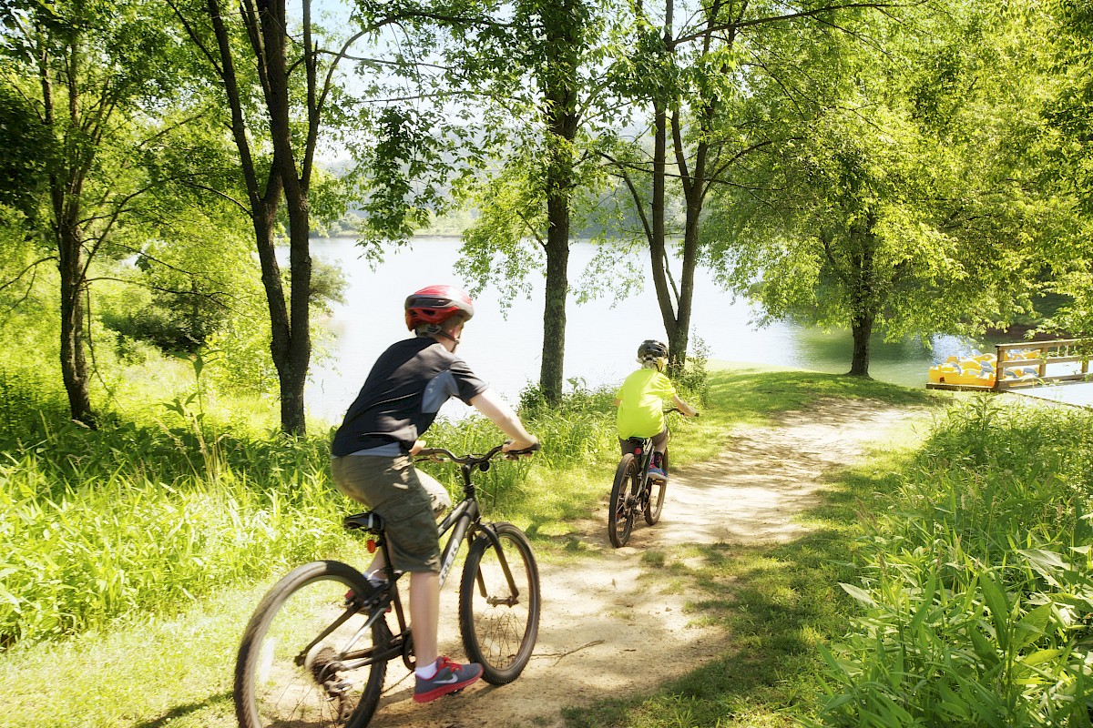 Two people riding bicycles on a dirt path through a green, wooded area near a body of water, wearing helmets, on a sunny day.