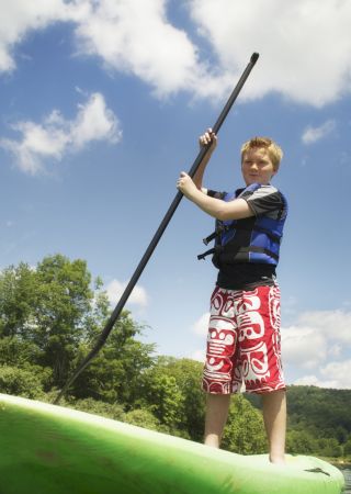 A boy wearing a life vest and red shorts is paddleboarding on a calm lake with lush greenery and hills in the background.