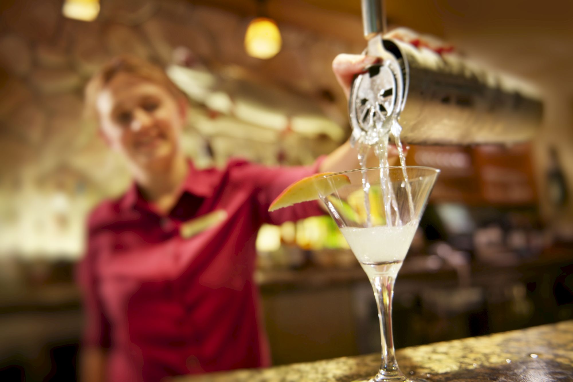 A bartender, wearing a red shirt, pours a cocktail from a shaker into a martini glass on a countertop, with a lemon wedge garnish.