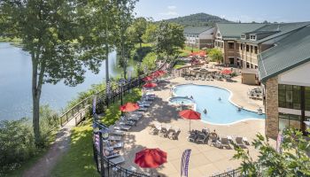 An outdoor pool area with red umbrellas, lounge chairs, and people relaxing, next to a building surrounded by greenery and a lake with mountains in view.