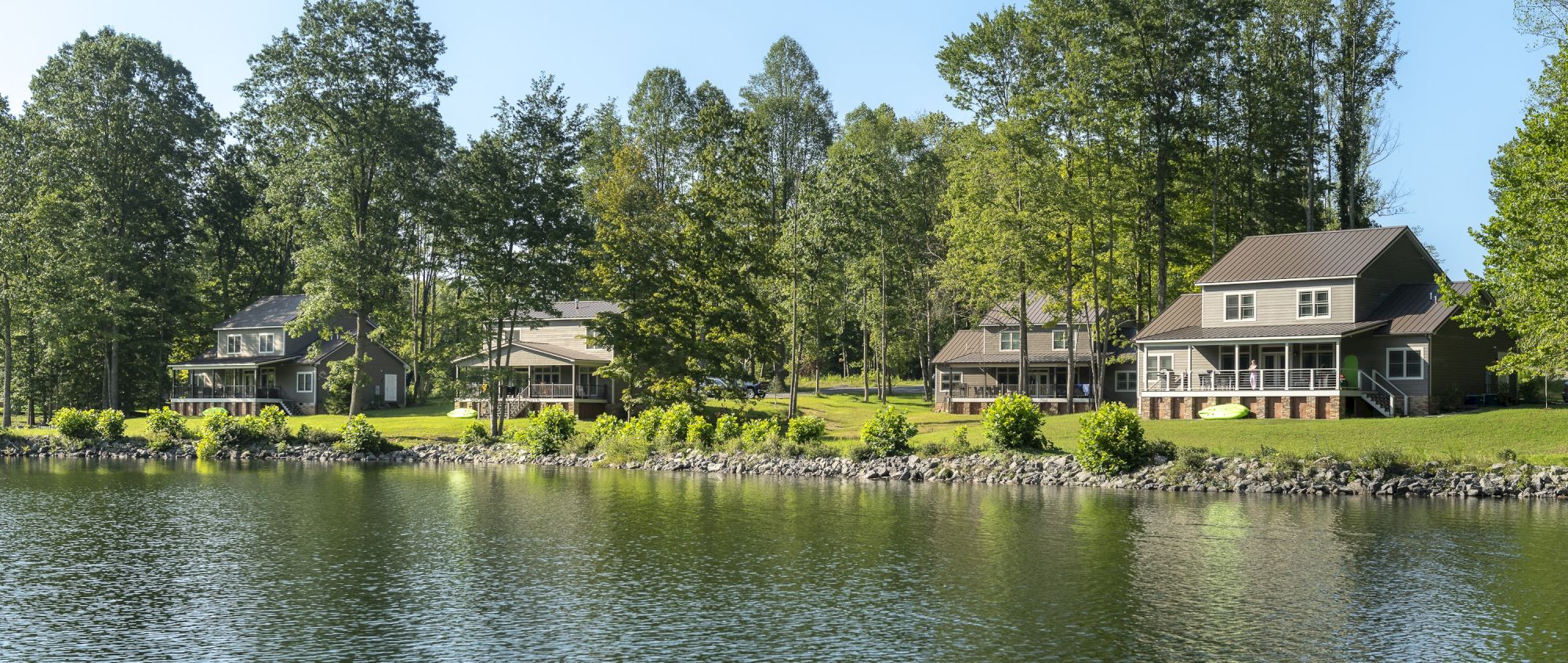 Several houses are situated along a tranquil lakeside, surrounded by lush trees and greenery, reflecting a peaceful and serene environment.