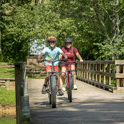 Two people are riding bicycles on a wooden bridge in a forested area. They are both wearing helmets.