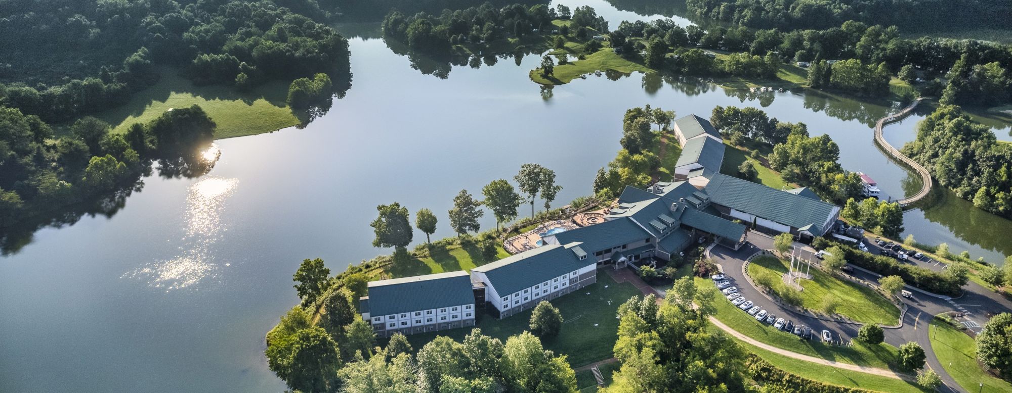 Aerial view of a building complex next to a lake surrounded by trees and greenery, with a reflective sun on the water ending the sentence.