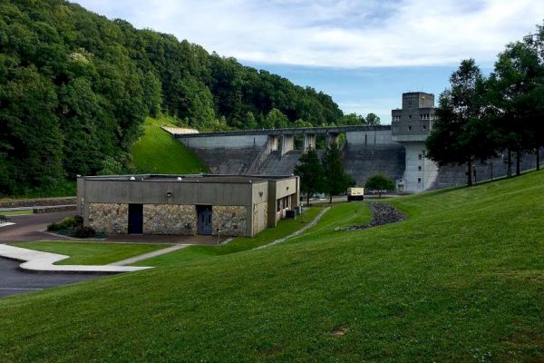 The image depicts a dam and its surrounding area, including a building, grassy hills, and trees, with a clear sky in the background, ending the sentence.