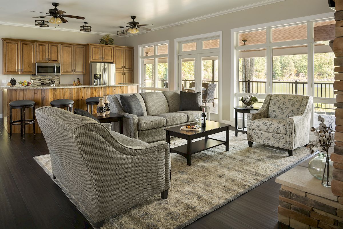 The image shows a modern open-concept living room and kitchen with grey furniture, wooden cabinets, bar stools, and large windows.