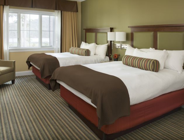 The image shows a hotel room with two neatly made beds, a window, a chair, and a nightstand with a lamp and phone. The room is well-lit and cozy.