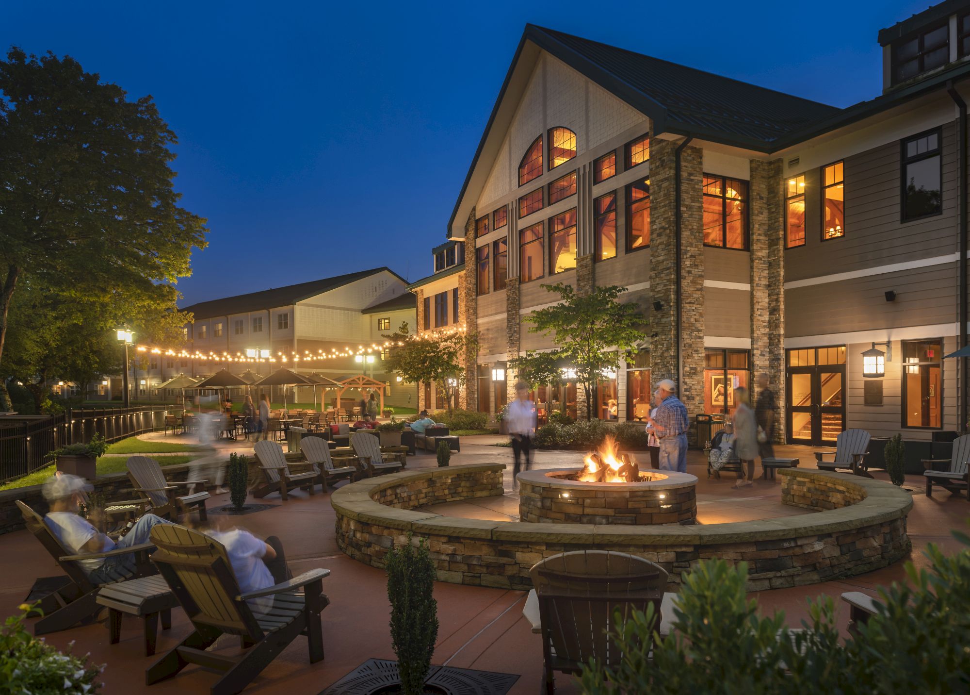 A cozy outdoor patio with a fire pit, surrounded by chairs, string lights, and a building in the background at dusk.