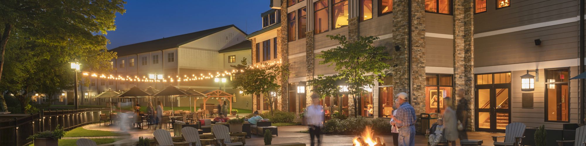 Outdoor area with a fire pit surrounded by chairs, string lights, people mingling, and a large building with lit windows in the evening.