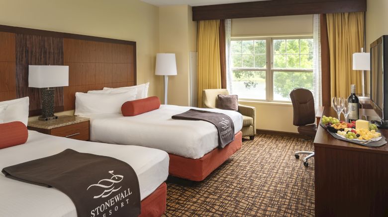 A hotel room with two beds, window, chairs, table, lamp, and a TV. Beds have linens labeled 