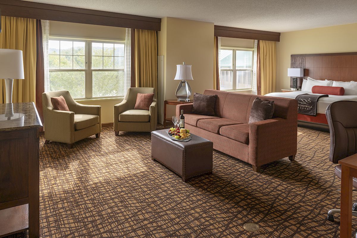 A hotel room features a bed, a sofa, a coffee table, two armchairs near a window, and two lamps, creating a cozy and inviting atmosphere.