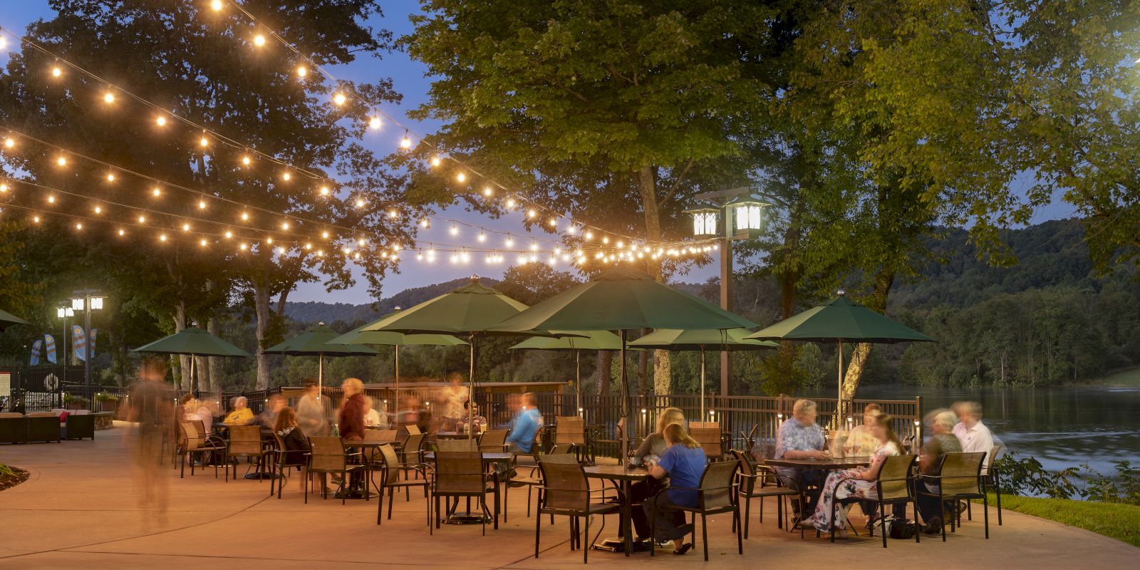 Outdoor dining area with string lights, green umbrellas, and people sitting at tables near a tree-lined waterfront during the evening.