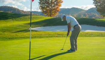 A person is putting on a golf green near a flagstick, with a sand trap and colorful trees in the background.