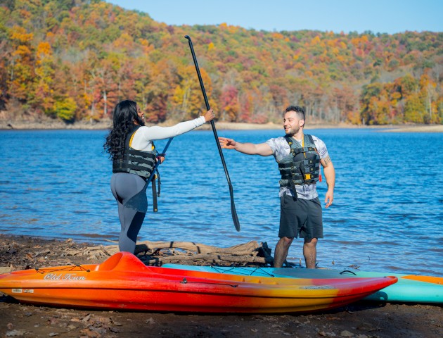 Two people wearing life jackets are holding a paddle beside kayaks on a lakeshore with autumn trees in the background.
