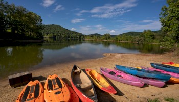 A serene lakeside setting with colorful kayaks and paddleboards on the sandy shore, surrounded by lush green trees and hills under a blue sky.