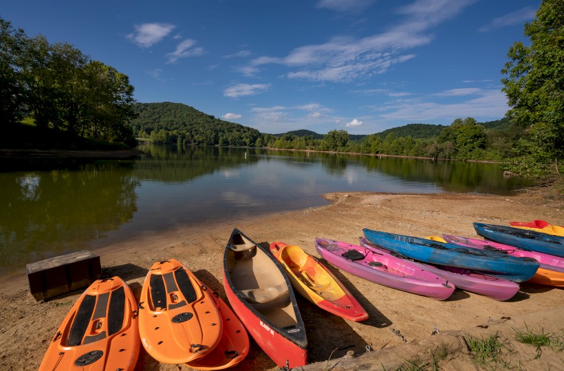 A serene lakeside setting with colorful kayaks and paddleboards on the sandy shore, surrounded by lush green trees and hills under a blue sky.