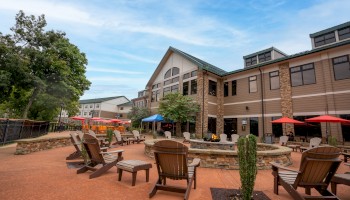 Outdoor patio area with wooden chairs, tables, and umbrellas in front of a multi-story building, surrounded by trees under a blue sky.