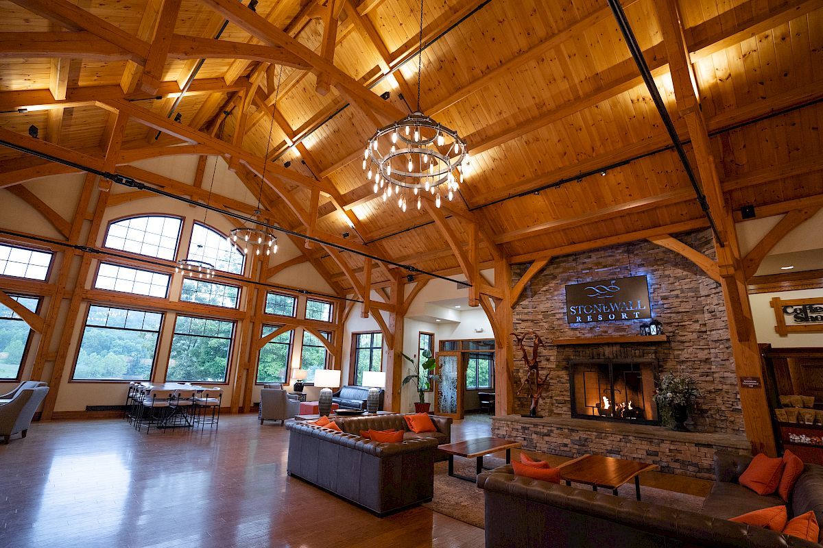 This image shows a spacious wooden lodge interior with high ceilings, large windows, cozy seating, a stone fireplace, and modern chandeliers.