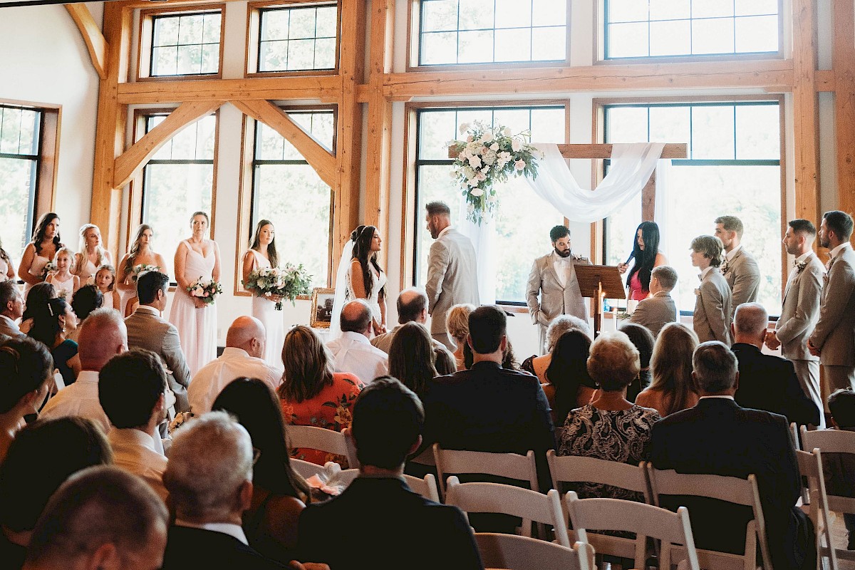 A wedding ceremony in a wooden chapel with guests seated, large windows, and a chandelier overhead, creating a warm, inviting atmosphere.